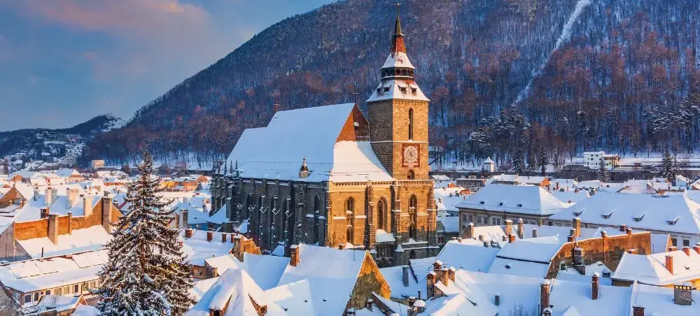 Snow-covered medieval town in Romania with Gothic church tower and traditional rooftops against mountain backdrop
