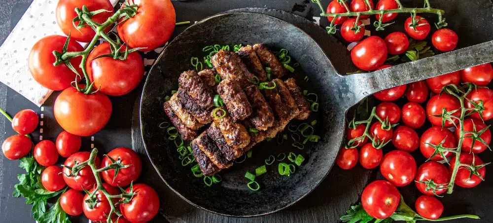 Grilled Romanian mici sausages in cast iron skillet surrounded by fresh vine tomatoes showcasing local cuisine