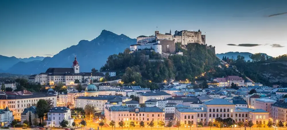 Historic Salzburg cityscape at twilight with illuminated baroque buildings and Hohensalzburg Fortress overlooking the Austrian Alps