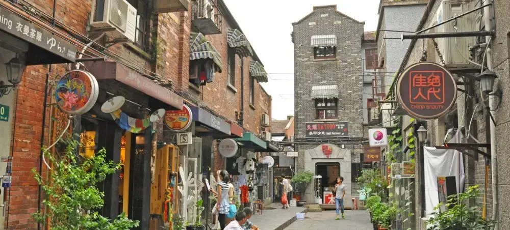 Narrow cobblestone alley in Shanghai's historic district with traditional shops, colorful signage, and visitors exploring local culture