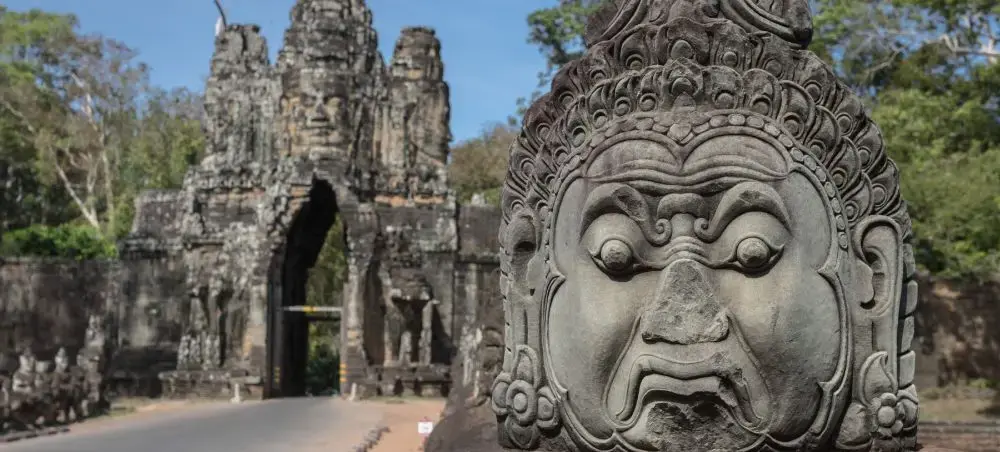 Ancient stone guardian statue at Angkor Thom entrance with Bayon temple towers in Siem Reap, Cambodia