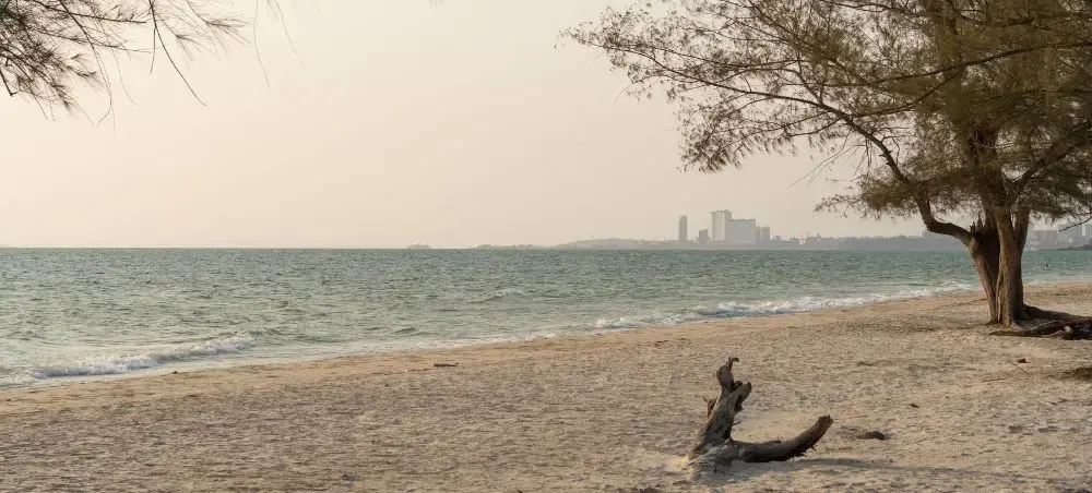 Pristine Sihanoukville beach with gentle waves, driftwood, and distant city skyline across calm waters