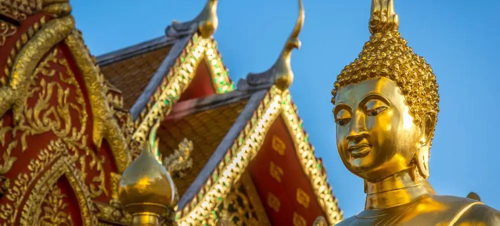 Golden Buddha statue with ornate Thai temple architecture featuring traditional red and gold decorative elements