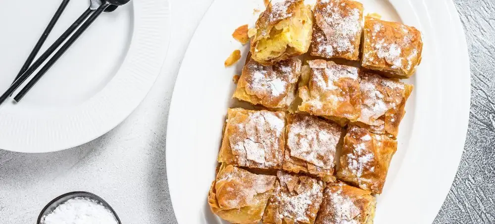 Golden bougatsa pastry squares dusted with powdered sugar served on white plate in Thessaloniki