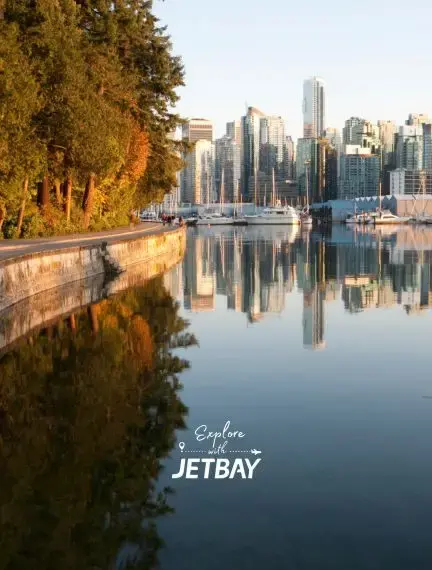Vancouver skyline reflecting in calm harbor waters with autumn trees along Stanley Park seawall