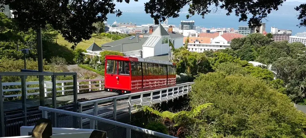 Red cable car ascending through lush Wellington hills with harbor and city skyline visible below