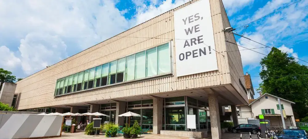 Modern Swiss airport terminal building in Zurich displaying "YES, WE ARE OPEN!" sign under bright blue sky