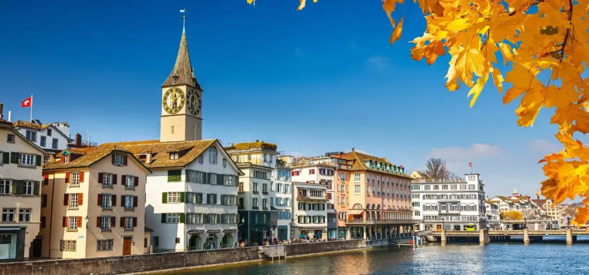 Historic Zurich waterfront with colorful medieval buildings, iconic clock tower, and autumn foliage framing the scene