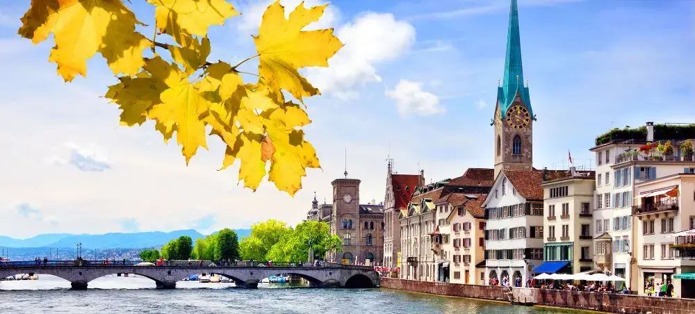 Zurich's historic waterfront with colorful buildings, church spire, and autumn foliage framing the scenic Swiss cityscape