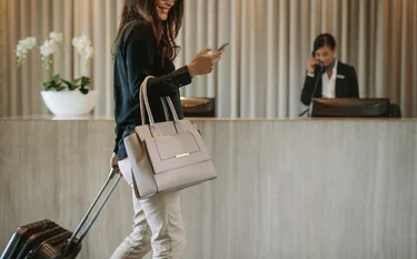 Business traveler with luggage checking phone at luxury hotel reception desk for private jet travel