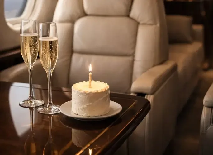Champagne flutes and birthday cake with lit candle on polished table inside luxury private jet cabin