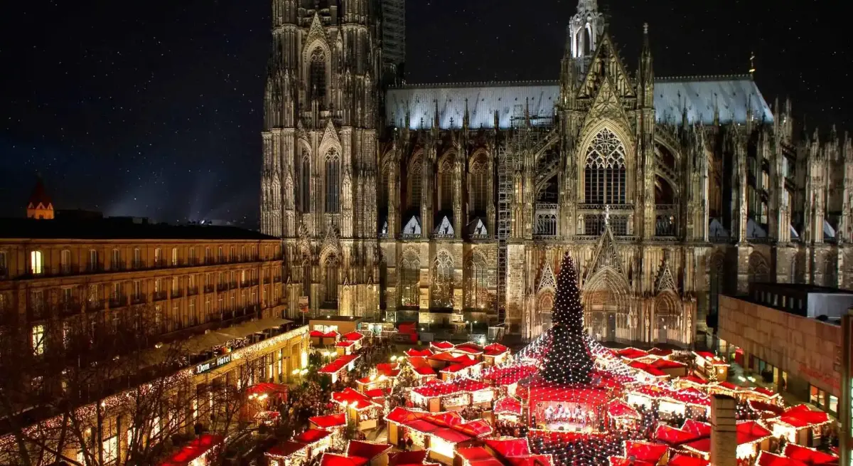 Cologne Christmas market with festive red-roofed stalls surrounding illuminated Gothic cathedral at night