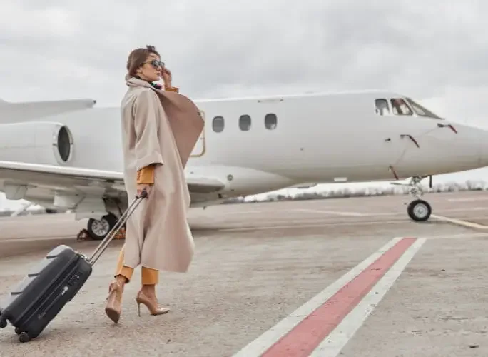 Elegant businesswoman in beige coat talking on phone while walking with luggage toward white private jet