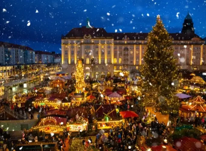 Festive Christmas market with illuminated stalls and towering tree beneath snowy evening sky in historic European city square