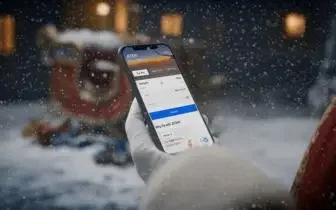 Hand holding smartphone displaying flight booking app with snowy Christmas scene and sleigh in background