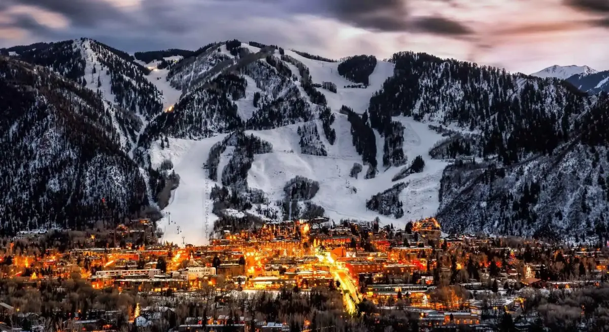 Illuminated alpine ski resort town nestled beneath snow-covered mountain slopes at twilight during winter season