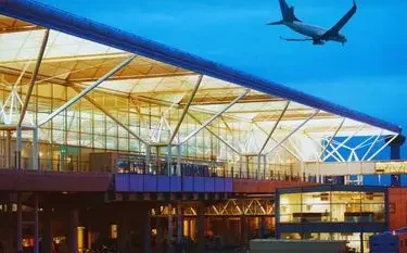 Modern London Stansted Airport terminal with glass architecture and aircraft overhead at dusk