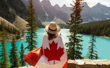 Person holding Canadian flag overlooking turquoise mountain lake surrounded by evergreen trees and peaks