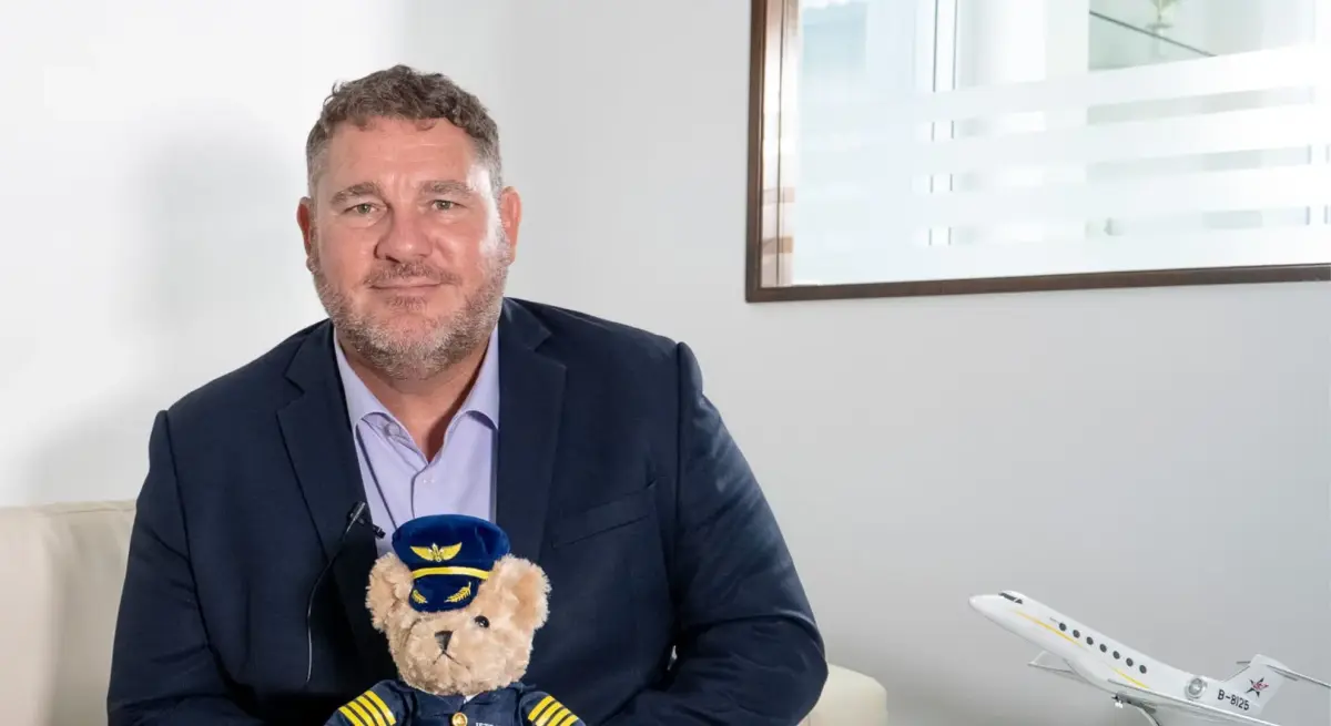 Phil Balmer in navy suit holding pilot teddy bear with private jet model, discussing Asian business aviation