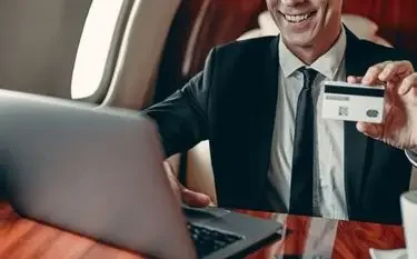 How Much Does a Private Jet Membership Cost? Smiling businessman using laptop and holding credit card aboard luxury private jet cabin