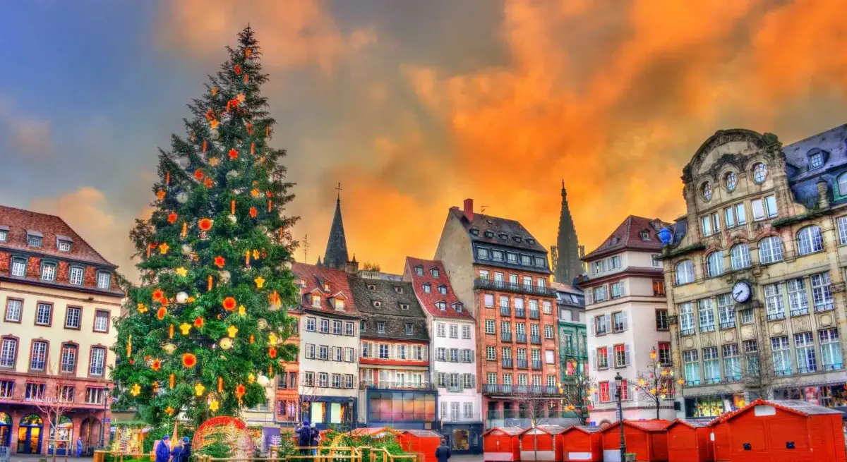 Strasbourg Christmas market with illuminated tree and festive red stalls beneath dramatic sunset sky