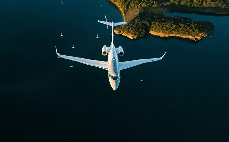 White private jet soaring above turquoise waters near rocky coastline with luxury yachts anchored below