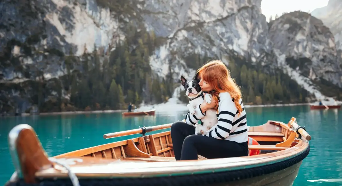 Woman cuddling Boston Terrier on wooden boat in alpine lake, showcasing luxury pet-friendly travel experiences