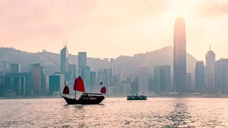Traditional red-sailed junk boat sailing past Hong Kong's iconic skyline at golden hour sunset