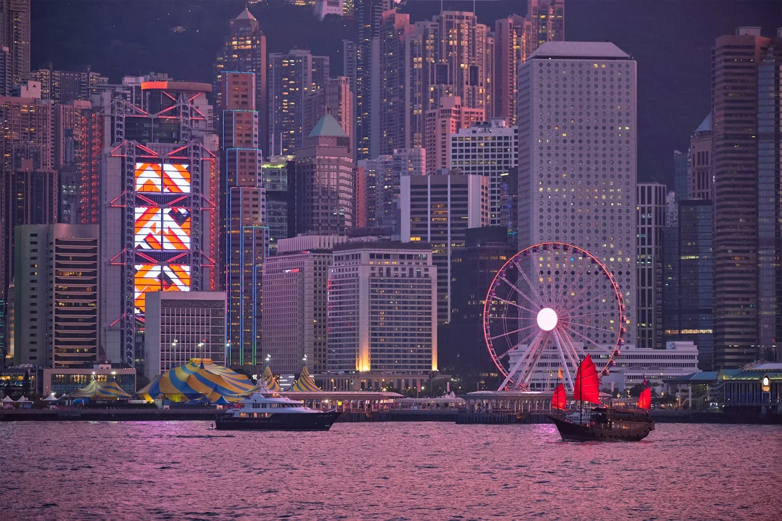 Hong Kong skyline at dusk with illuminated ferris wheel and traditional red-sailed junk boat on Victoria Harbour