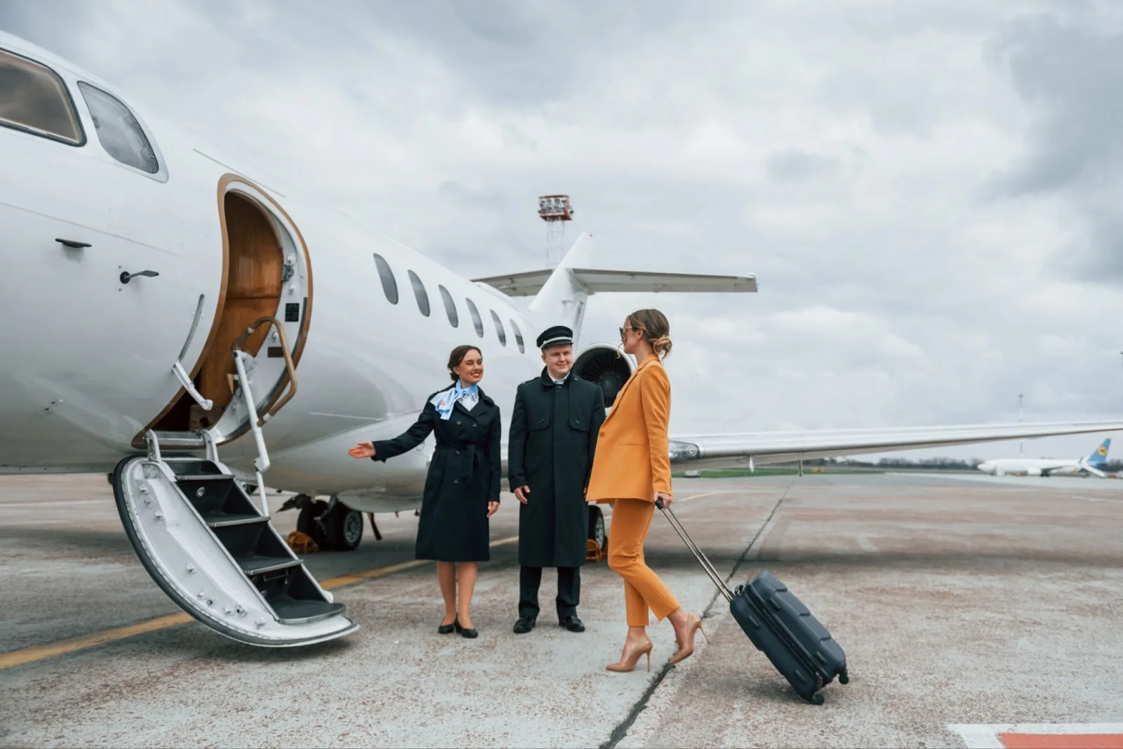 Professional flight crew welcoming passenger boarding white private jet with luggage on airport tarmac