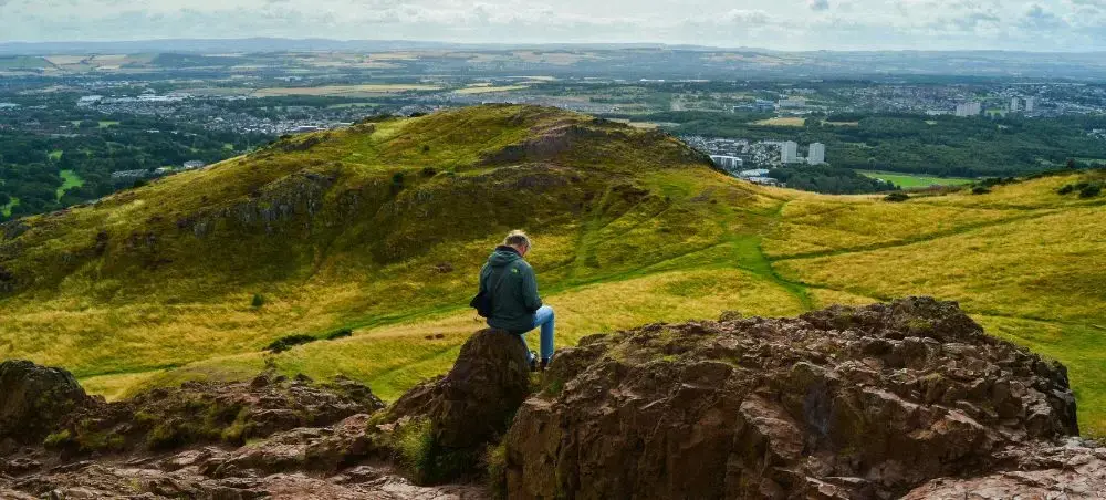 Traveller sitting on rocky summit of Arthur's Seat overlooking Edinburgh's rolling hills and cityscape beyond