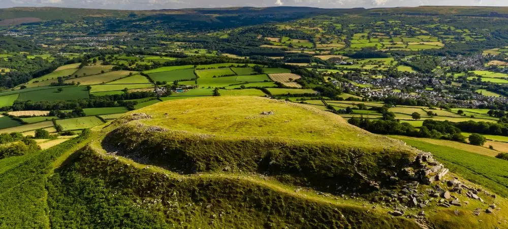 Aerial view of a grassy hilltop summit with rocky outcrops overlooking the lush patchwork valleys and market towns of Bannau Brycheiniog, Wales