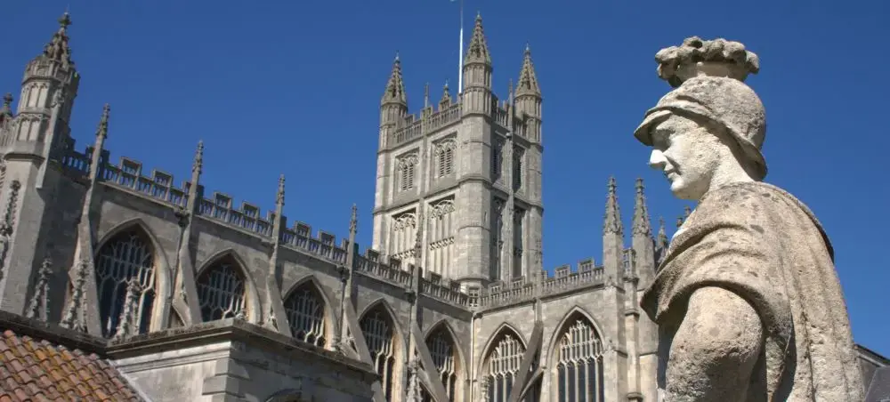 Stone Roman soldier statue with Bath Abbey's Gothic towers and intricate fan vaulting rising against a clear blue sky