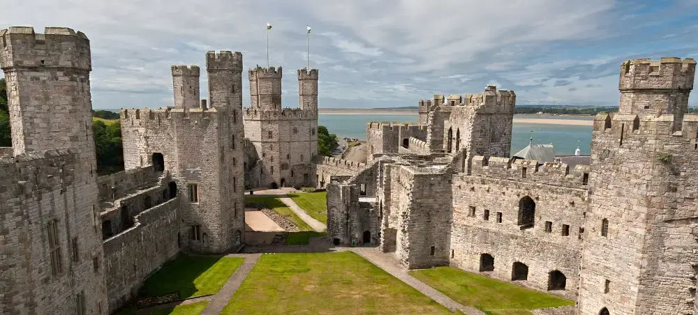 Aerial view of Caernarfon Castle's medieval stone towers and battlements with the Menai Strait and North Wales coastline beyond