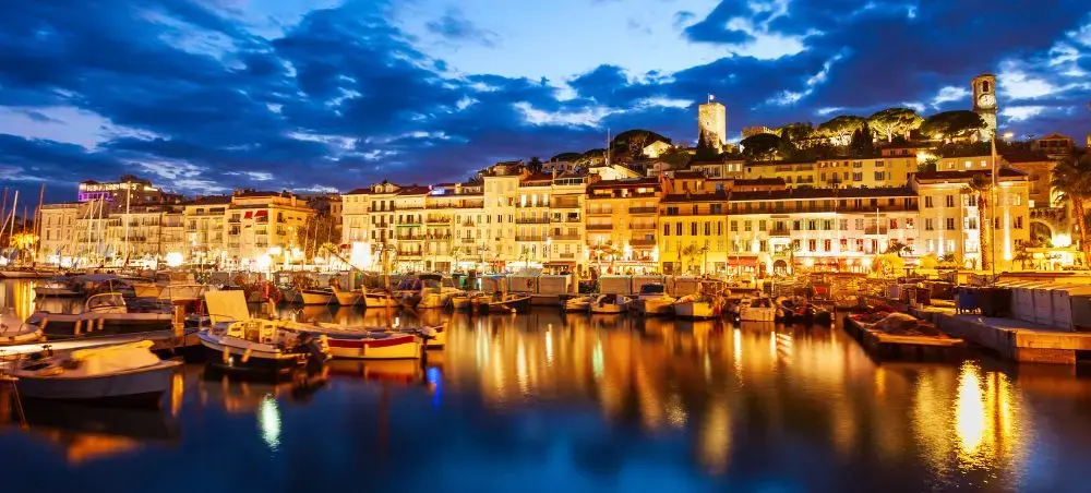 Cannes Old Port illuminated at dusk with luxury yachts moored along the waterfront and the historic Le Suquet hill glowing against a dramatic blue sky