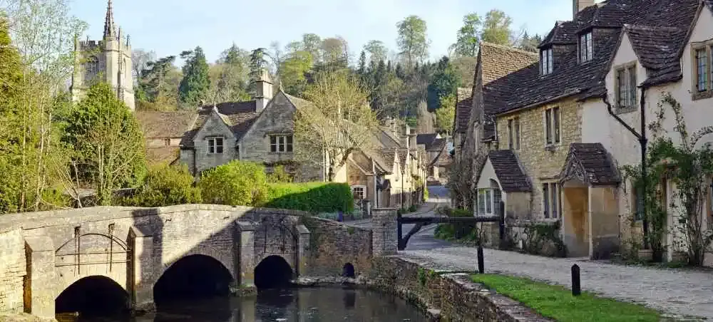 Cotswolds village with cottages, a medieval stone bridge over a stream, and a church spire rising above spring trees