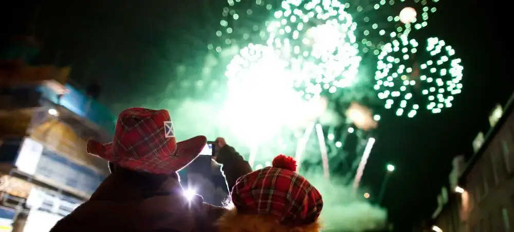 Couple in Scottish tartan hats watching green fireworks light up the Edinburgh night sky during Hogmanay celebrations