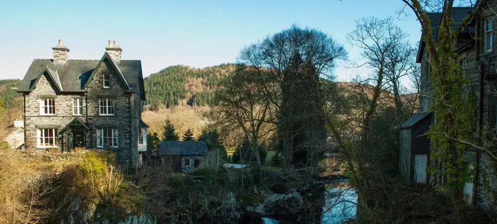 Stone country houses beside a stream in Eryri, Snowdonia, with forested hills and clear blue sky