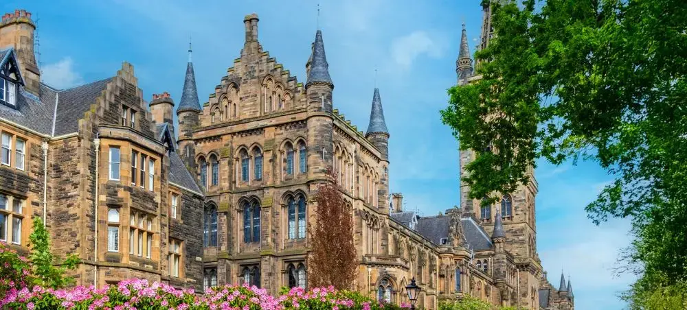 Gothic sandstone architecture of Glasgow University with ornate spires and pink flowering shrubs under a blue sky