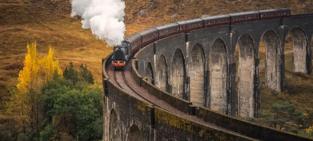 Steam train crossing the Glenfinnan Viaduct in the Scottish Highlands, surrounded by autumn foliage