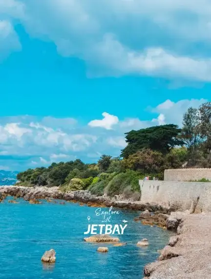 Crystal-clear turquoise waters along the rocky shoreline of Îles de Lérins near Cannes, framed by lush Mediterranean pines and a historic stone wall under a bright blue sky