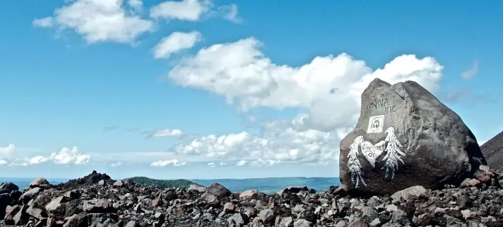 Volcanic rock summit with painted angel wings graffiti overlooking a rugged lava field landscape under blue skies from Cerro Negro volcano