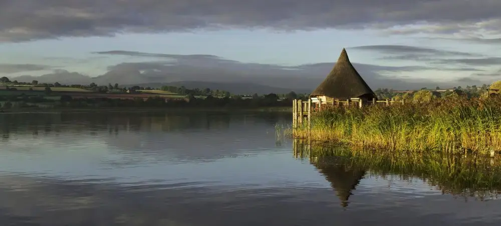 Thatched crannog-style boathouse reflected in the still waters of Llangorse Lake, Bannau Brycheiniog, with rolling Welsh hills and dramatic clouds at dusk