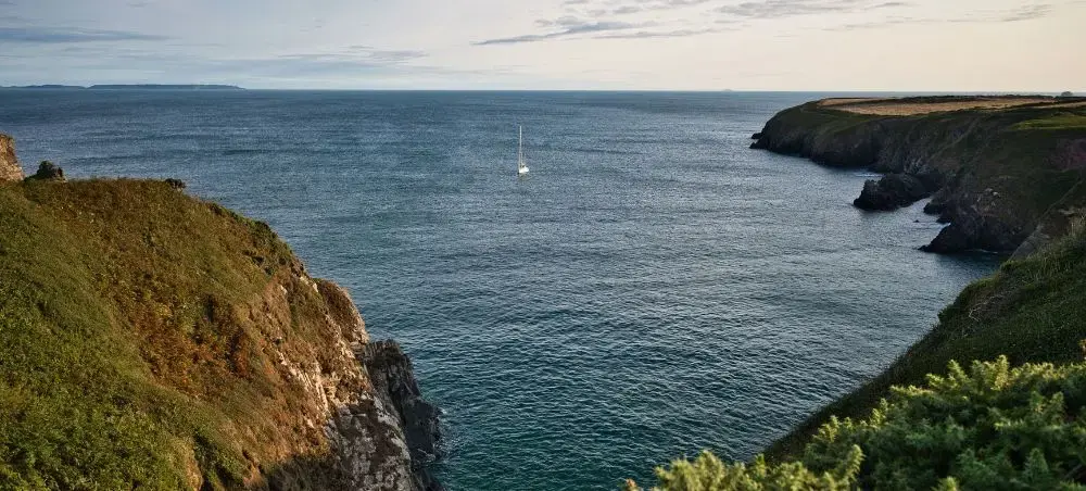 Dramatic Pembrokeshire coastline with rugged cliffs, lush green headlands, and a sailboat on calm waters off West Wales
