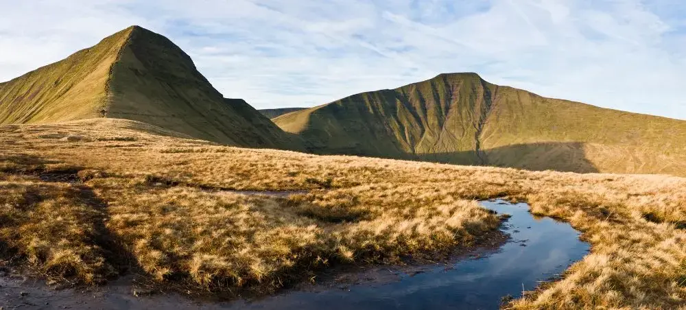 Panoramic view of Pen y Fan and Corn Du peaks in Bannau Brycheiniog, with golden moorland grasses and a reflective mountain pool under blue skies