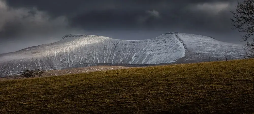Snow-dusted twin peaks of Pen y Fan and Corn Du rising dramatically under stormy skies in Bannau Brycheiniog, South Wales