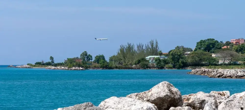 Jet aircraft on final approach over the turquoise Caribbean Sea near Montego Bay, Jamaica, with lush tropical coastline and rocky shoreline in the foreground