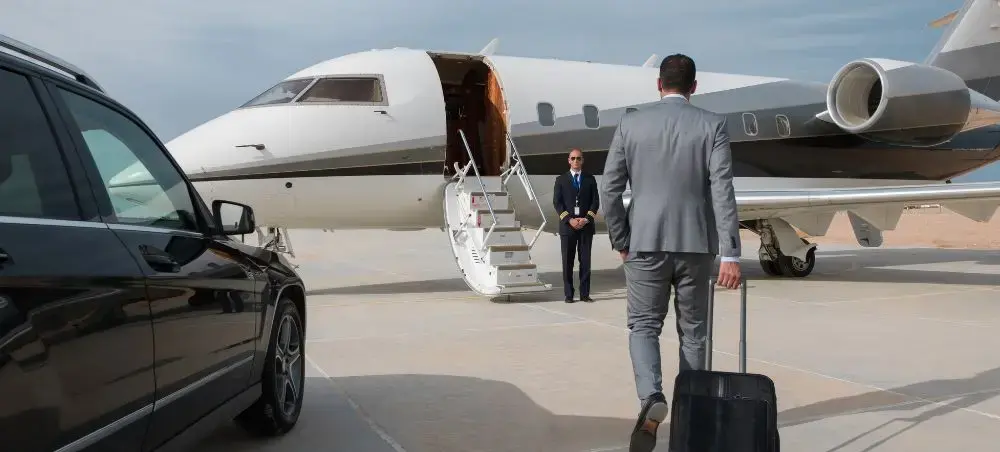 Business traveller with luggage walking across the tarmac to board a private jet charter, greeted by a pilot at the steps