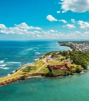 Aerial view of Puerto Plata coastline featuring historic fort, lush greenery, and turquoise Caribbean waters on a clear sunny day