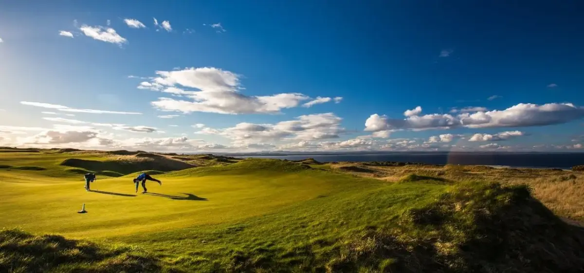 Golfers reading the green on a dramatic links course overlooking the sea near St Andrews, Scotland, under a vivid blue sky